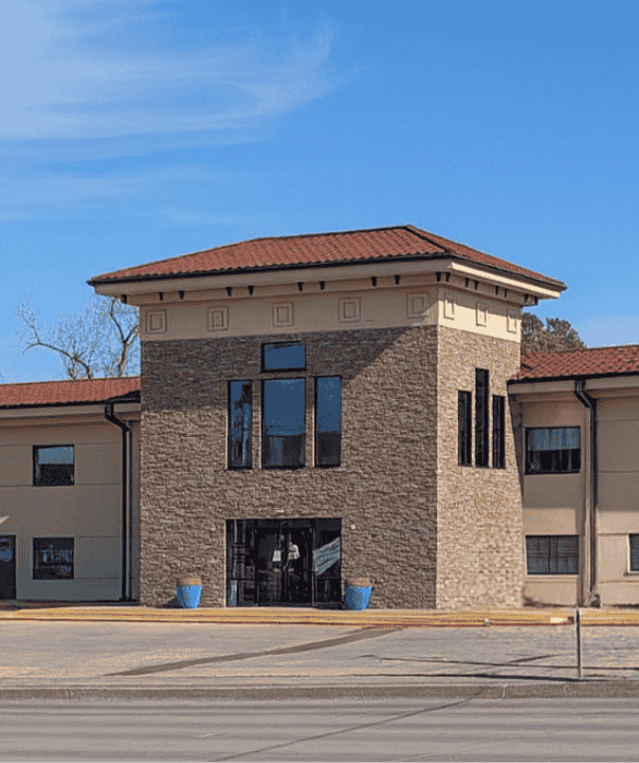 A modern two‑story clinic with tan walls, stone accents, and large windows stands beneath a clear blue sky, welcoming patients to AAIC.