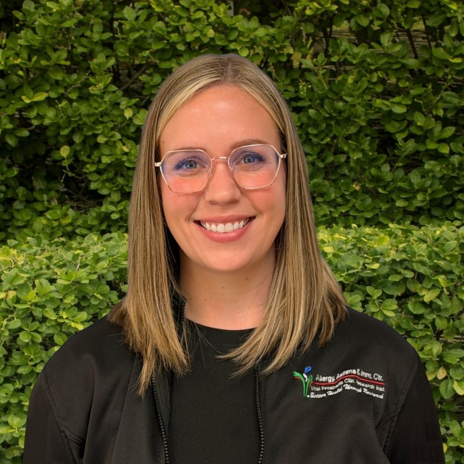 A member of the staff in a black jacket stands before green foliage, representing welcoming patient support within the allergy and asthma clinic.