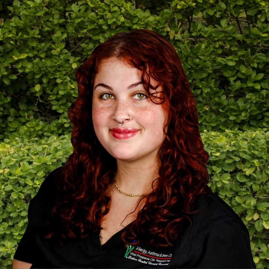 A clinical staff member with curly red hair stands outdoors in AAIC scrubs, reflecting supportive patient care in allergy and immunology.