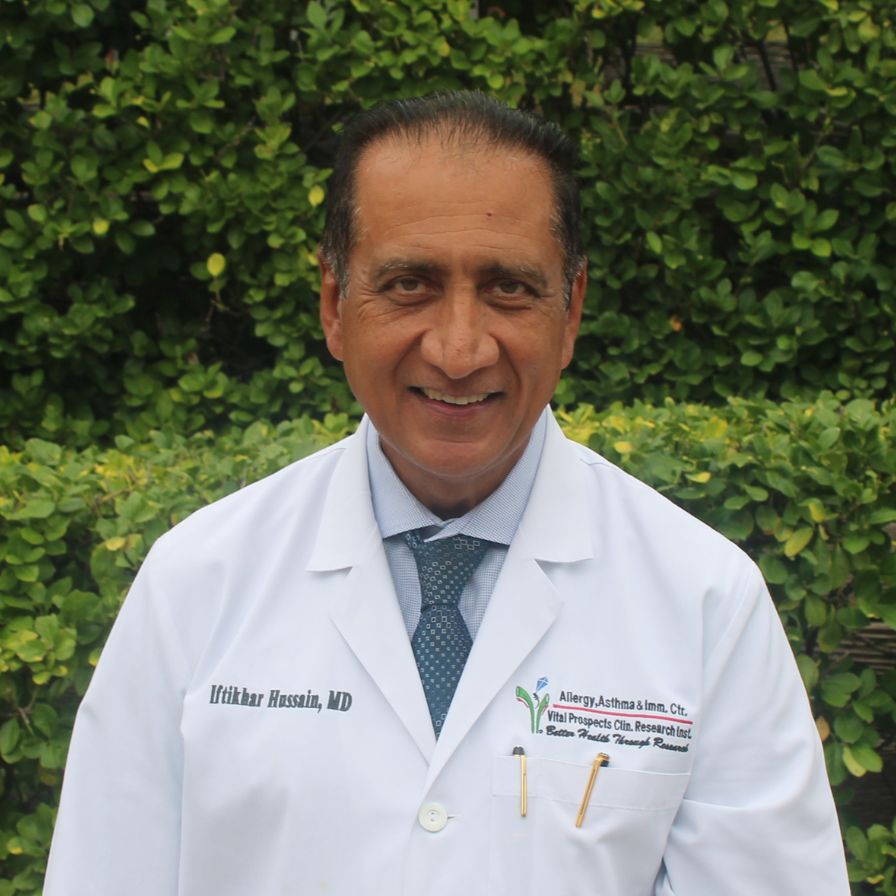 A physician in a white lab coat and tie stands in front of greenery, reflecting clinical leadership in allergy, asthma, and immunology services.