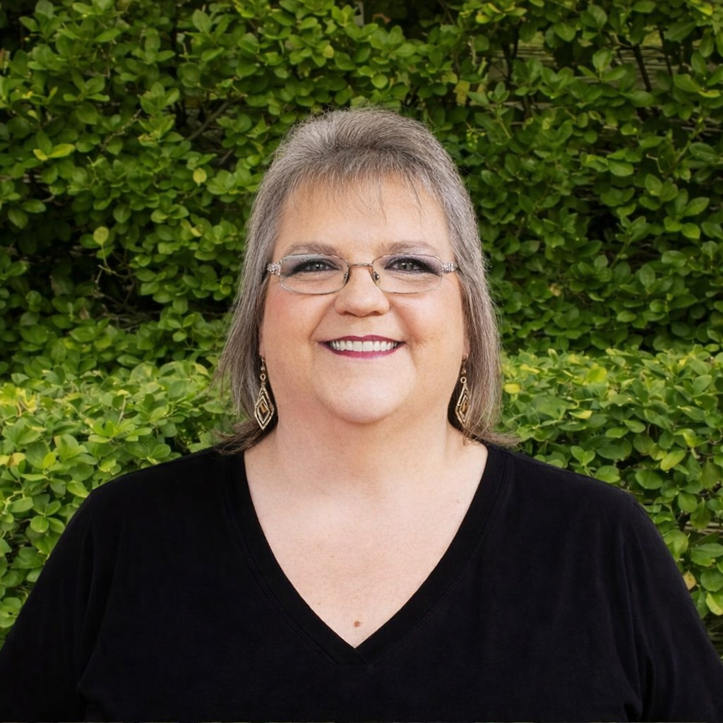 A smiling staff member a black shirt stands before green foliage, representing patient support within the allergy and asthma clinic.