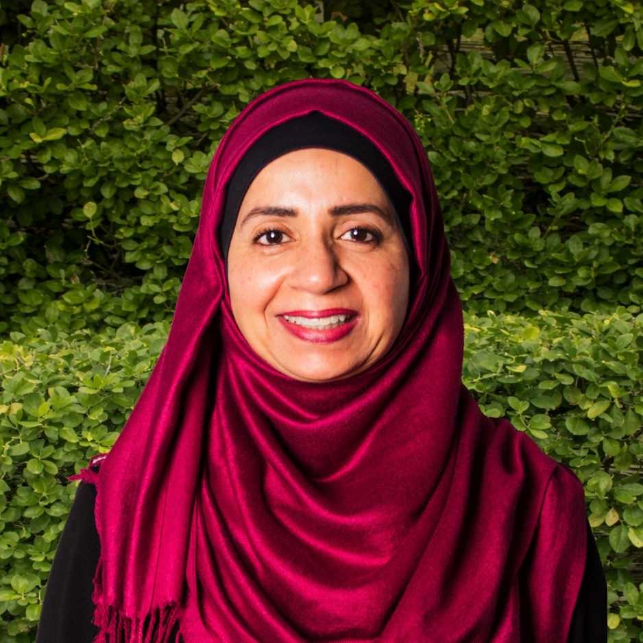 A team member wearing a deep red hijab stands outdoors against greenery, reflecting dedicated support in allergy and immunology services.