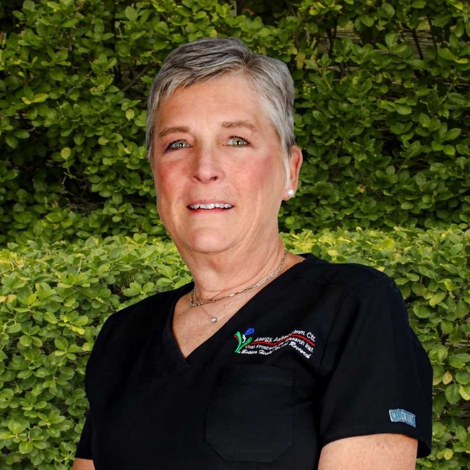 A medical staff member in black scrubs stands outdoors before green foliage, reflecting professional support in allergy and immunology care.
